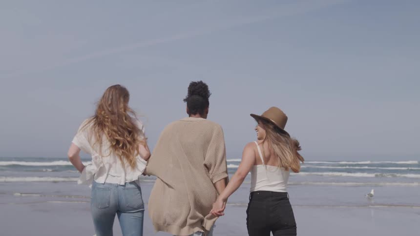 Female friends holding hands running and laughing towards the shoreline at the beach