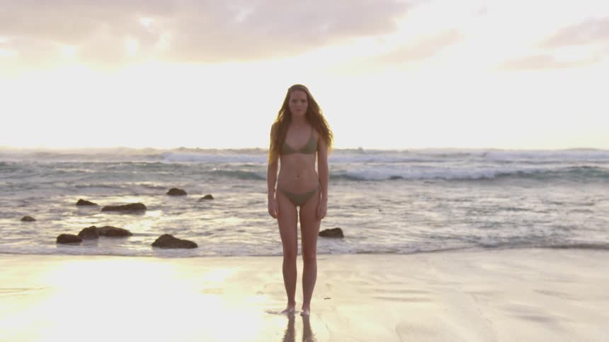 A woman stands in a two piece bikini on the beach with her arms by her side and looks into the camera