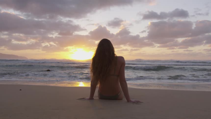 Woman wearing a bikini and sitting on the sand beach looks at the sunset next to the ocean