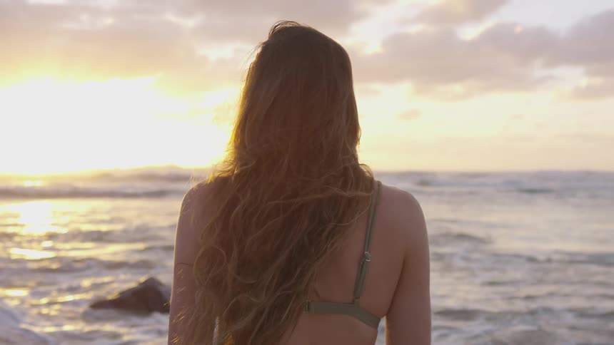 A woman in a two piece bikini stands on the beach at sunset