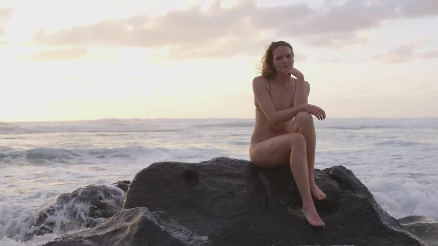 A white woman wearing a bathing suit sits on a rock at the beach