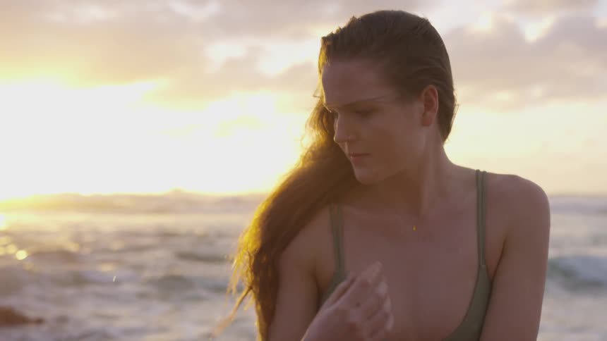 Blonde woman wearing a swimsuit touches her hair on the beach at sunset next to the ocean