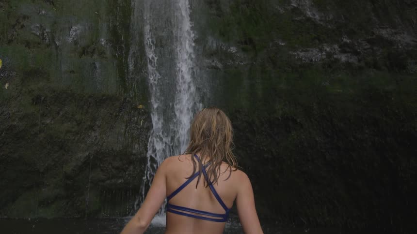 A woman in a swimsuit stands in front of a waterfall flowing down a mountain side