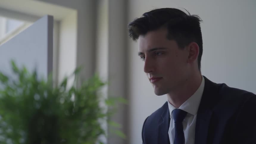 A man in a suit standing up staring off into the distance with a plant in the background