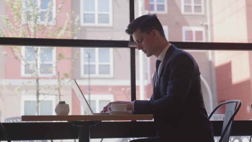 White businessman in a blue suit sitting at the table uses a laptop and sips on a cup of coffee in the cafe