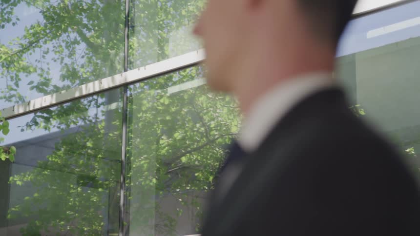 A man in a dark suit stands outside of a building upon which the reflection of a tree can be seen