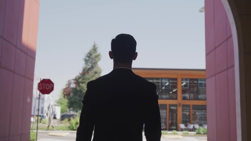 A man walks under an archway and out to a parking lot on a sunny day