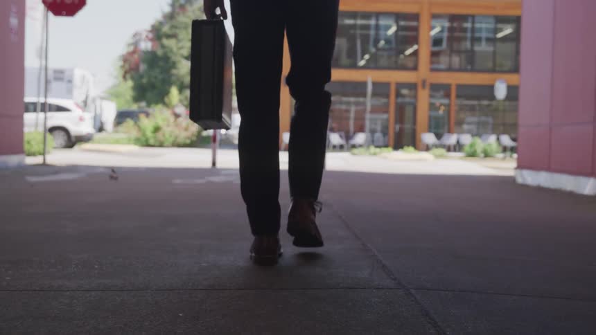 A man carrying a briefcase is walking beneath an overpass towards a street and building in front of him