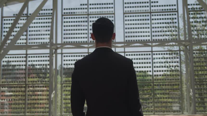 A man stands and looks out a window at the building across from him