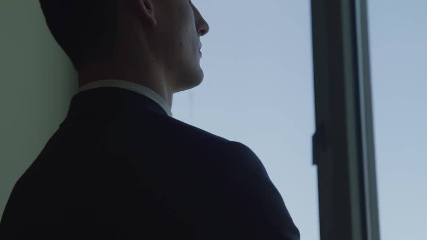 A young man stands in his office and looks out the window