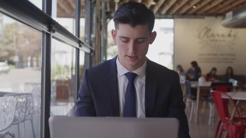 A man wearing a suit is typing on a laptop while in a cafe