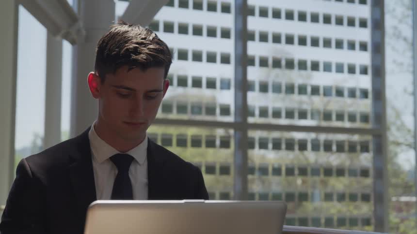 A man sits at a desk with a laptop and large windows behind him