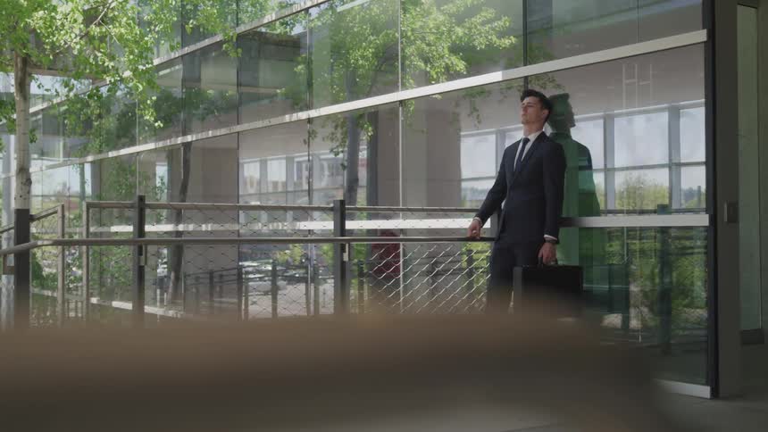 Business man wearing a suit and holding a suitcase leans against window in front of the office building