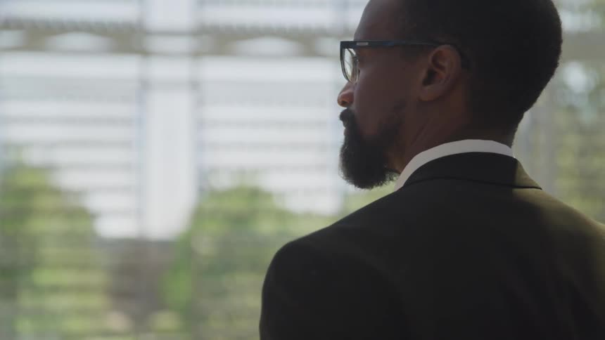 A man in a business suit stands looking out atrium windows