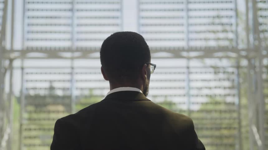 A black male in a suit coat stands before a scaffold with a view of the outdoors