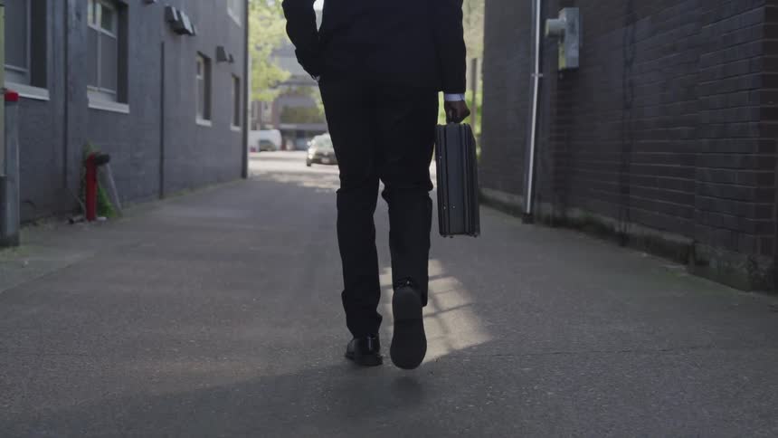 Businessman wearing a suit and carrying a suitcase walks along the road between two buildings