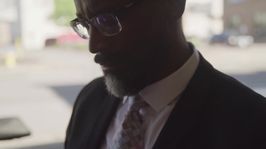 A black man in a suit standing outdoors in shadow of a building in the city during the day