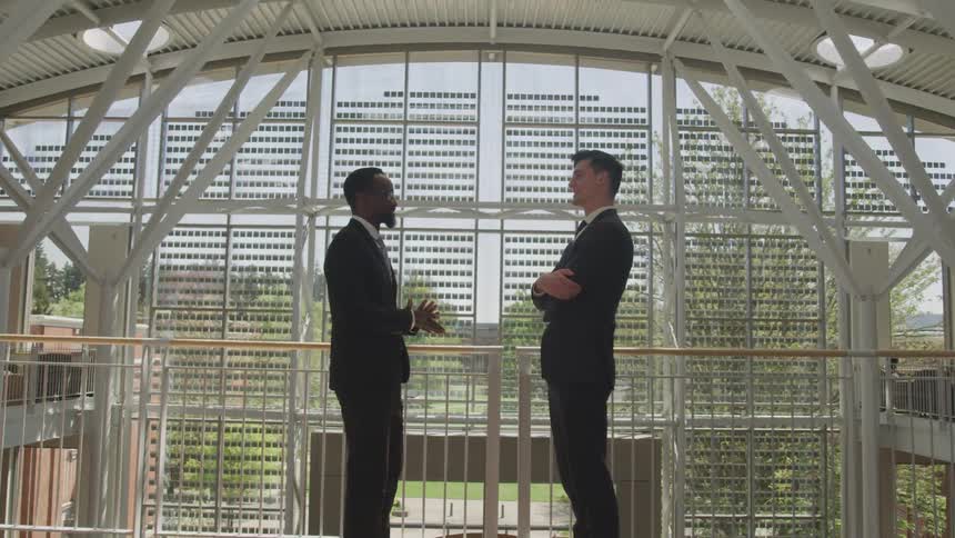 Two men insuits stand facing each other talking near a railing in a building atrium