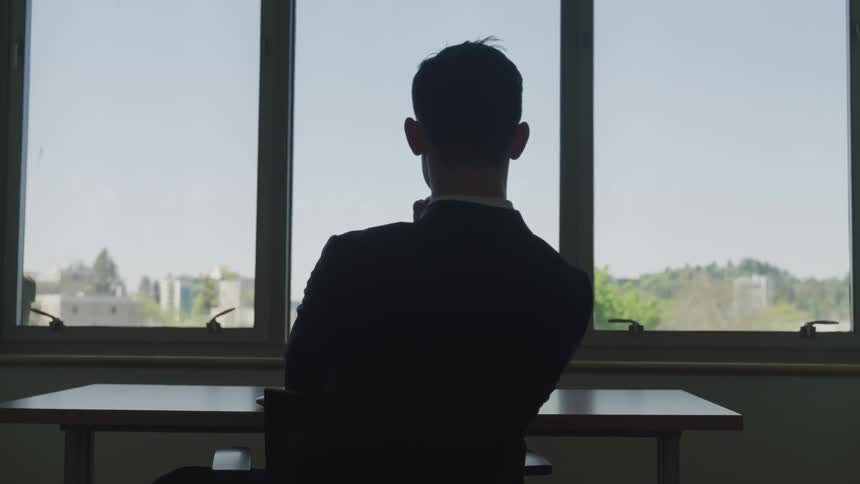 A man sits at a desk in front of a window and gazes outside