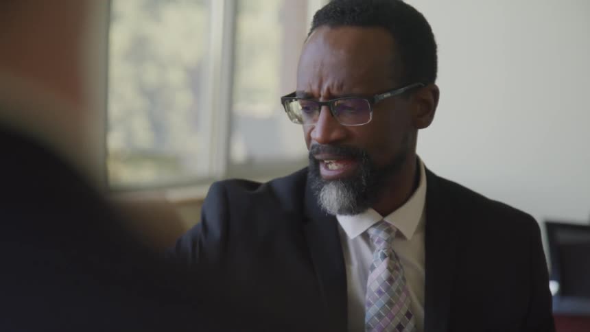 Black man sits at a table near a window as he talks to another man