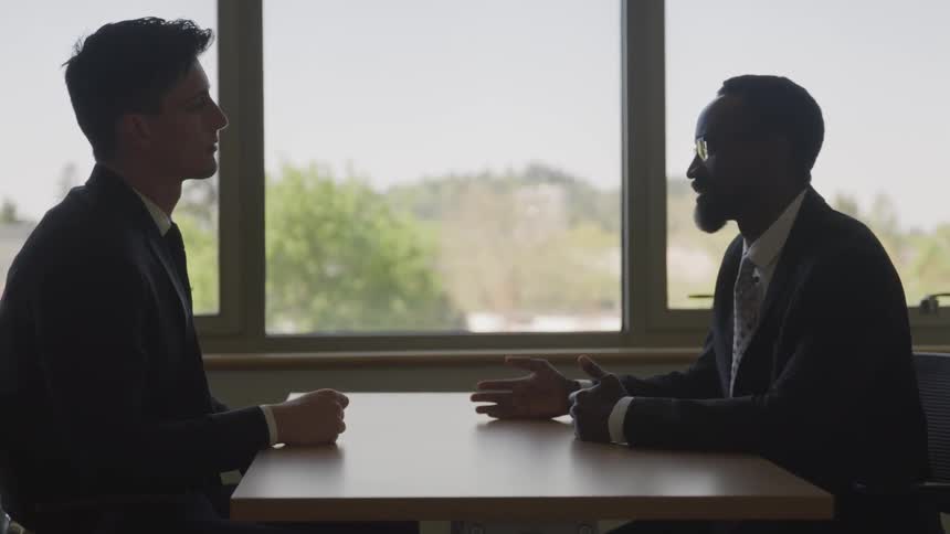 Two men in suits sit at a table and talk