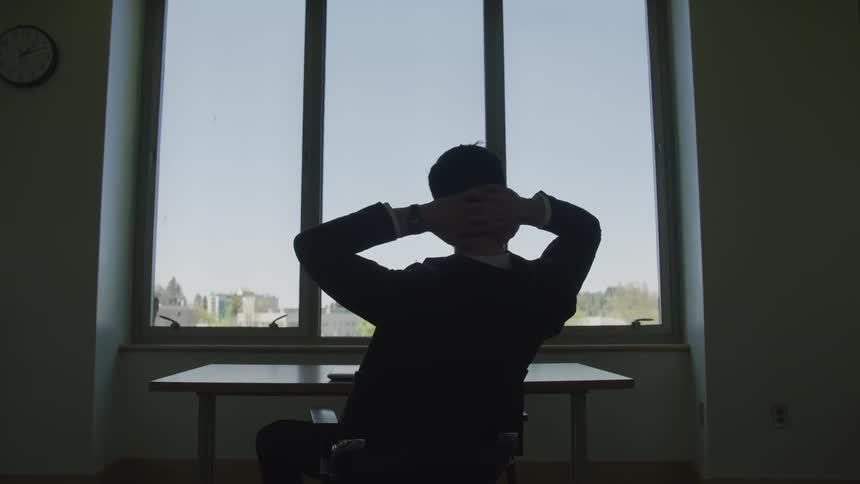 A man leaning back in his chair with his arms behind his head looking out a window