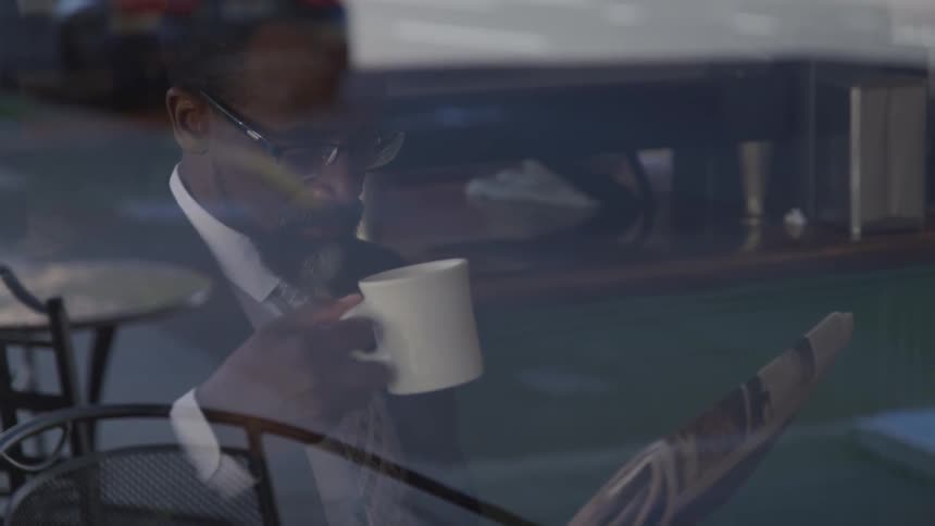 A black man drinks a cup of coffee while reading the newspaper