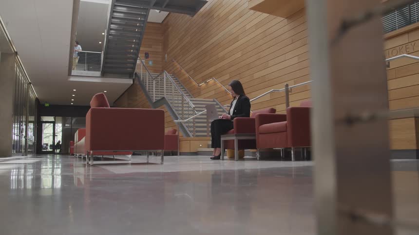 A woman sits in a chair in a lobby typing on a laptop