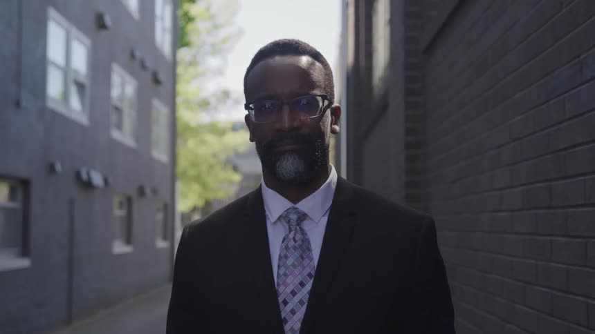 Black businessman wearing a suit and glasses stands between two buildings in the street