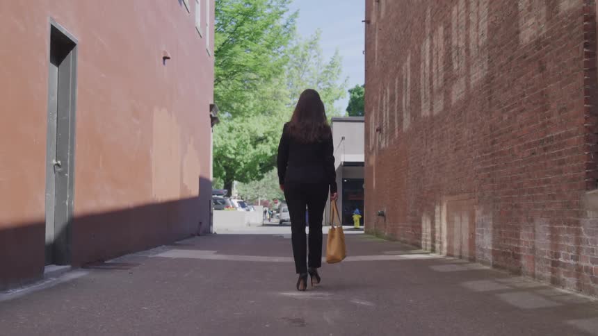 A woman in a suit and heels carrying a bag walks down an alley on a sunny day