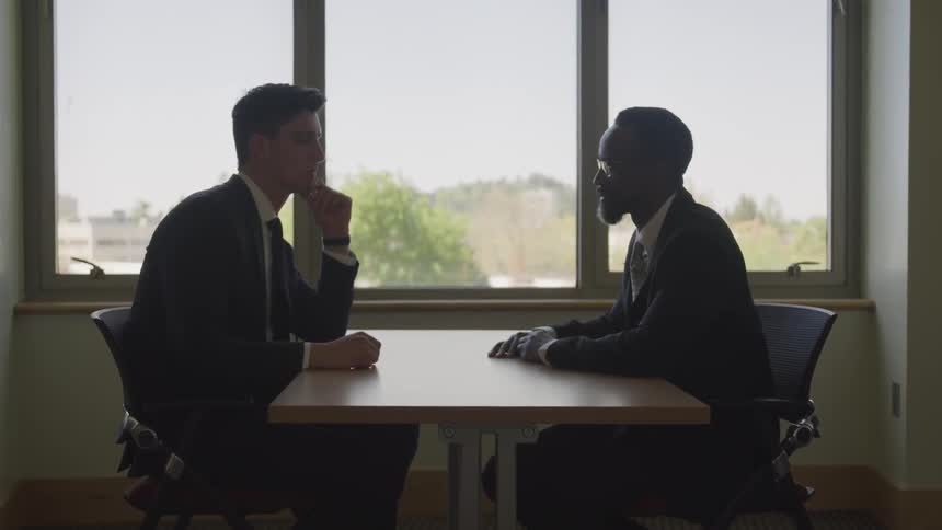Two businessmen face each other across table next to window
