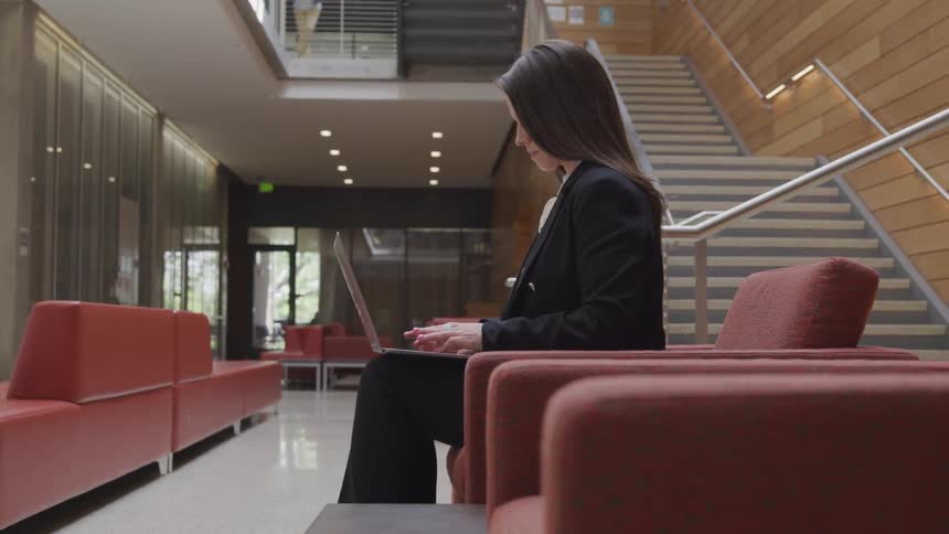 A woman sitting up straight in a building reception area types on a laptop