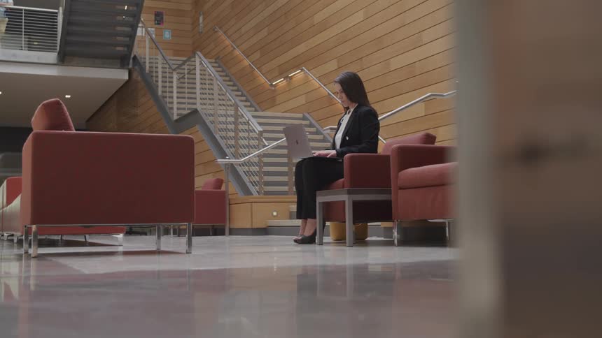 Female sitting on a chair in a lobby typing on a laptop that is balancing on top of her legs