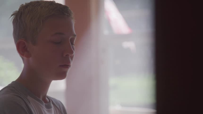 A boy inside staring off at the distance with a window in the background