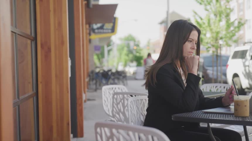 A woman sits at an outdoor patio restaurant table with a drink and laptop