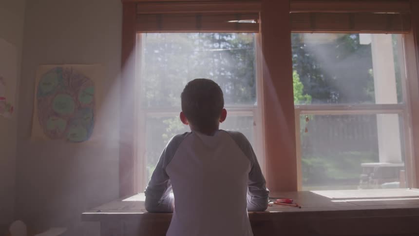 A young male sits at table in front of a window through which sunlight is pouring in