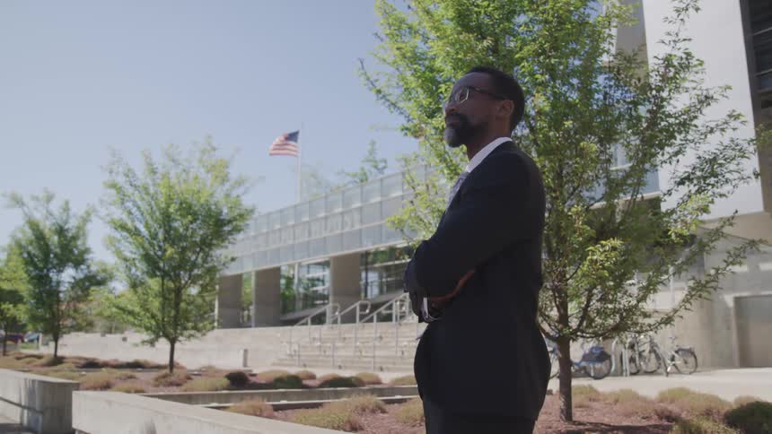 A middle aged man stands in front of a building with an american flag waving on top of it