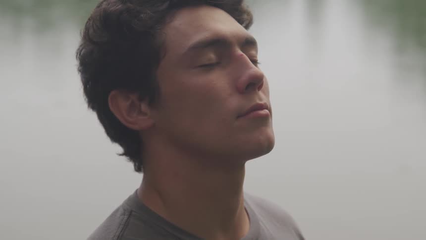 Man with wavy hair meditating while standing next to a calm lake