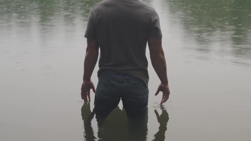 A man is standing in calm water up to his thighs while it sprinkles