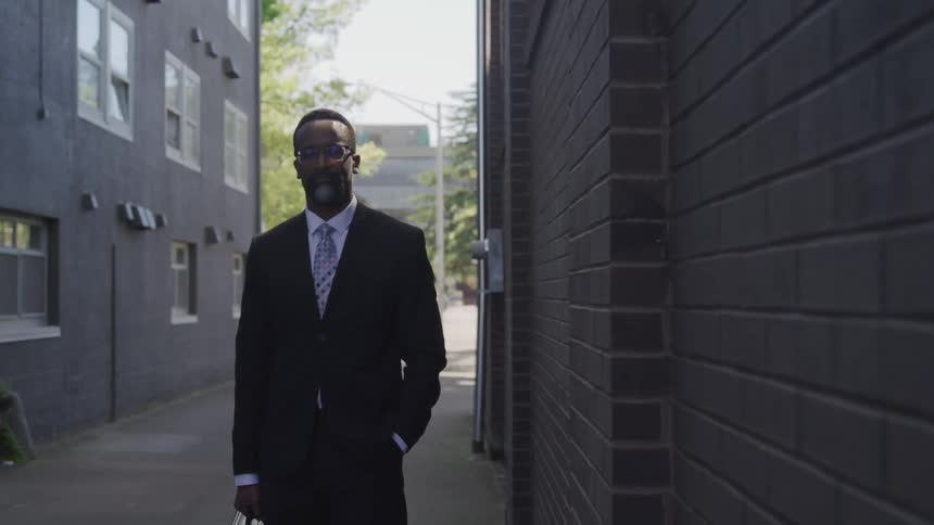 A man in a suit carrying a briefcase stands in a city alley between two buildings