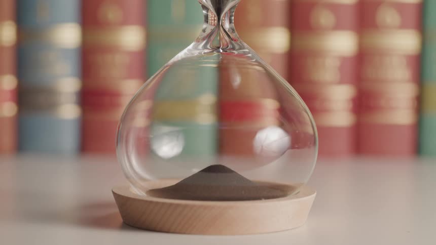 An hourglass with brown sand with books in the background