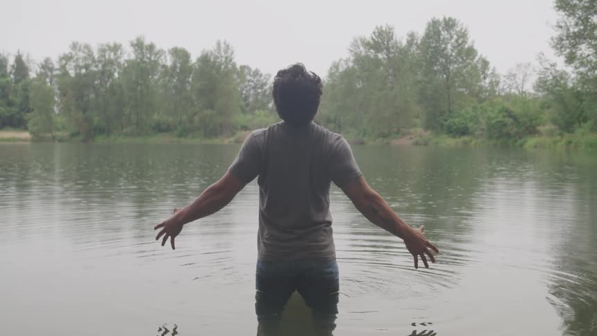 A man stands in the water and raises his arms up and looks up to the sky