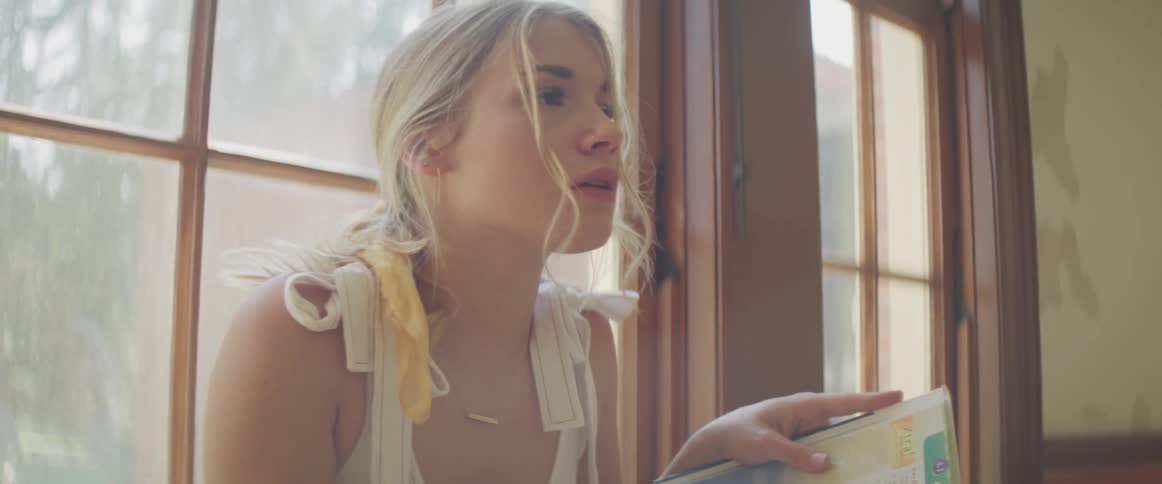 A girl sits on the window ledge of a room writing in a binder after looking up and thinking