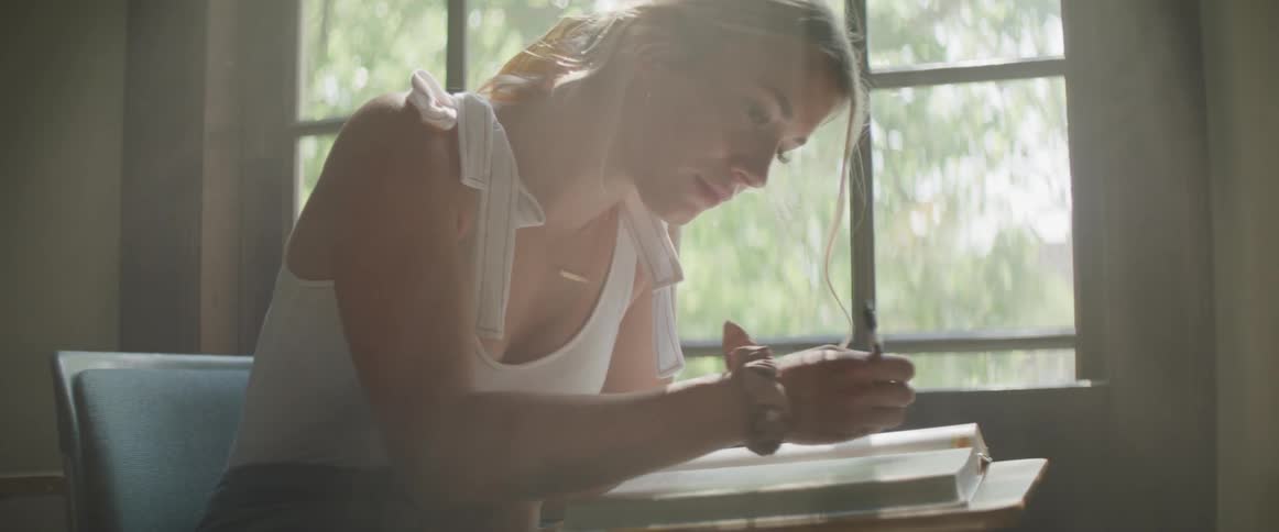 A young woman is seated at a desk reading a book and writing notes