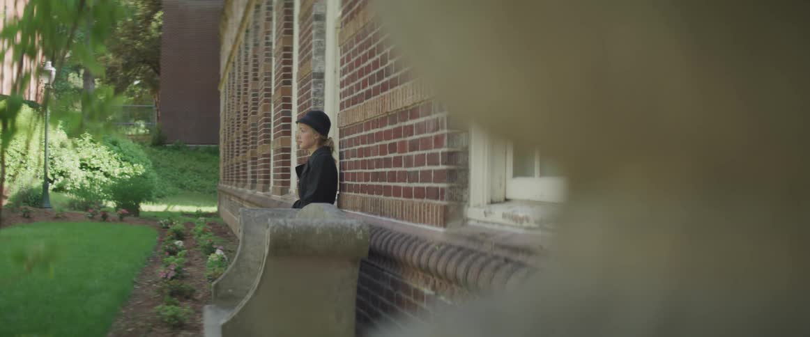 Blonde woman wearing a black hat and coat leans against brick wall of a building