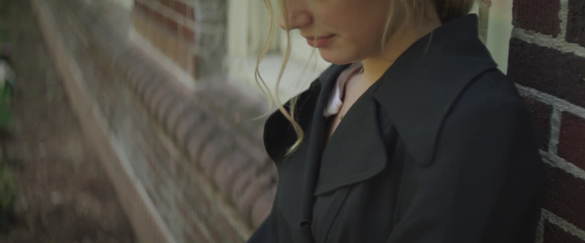 Blonde woman wearing a black coat leans against the brick wall of the building