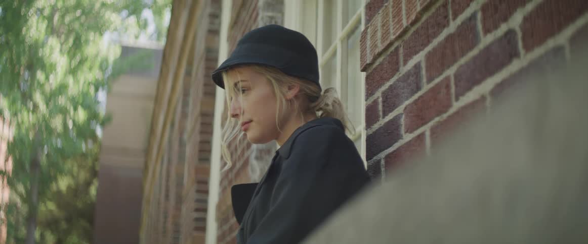 A young woman leans against a brick wall and looks ahead of her