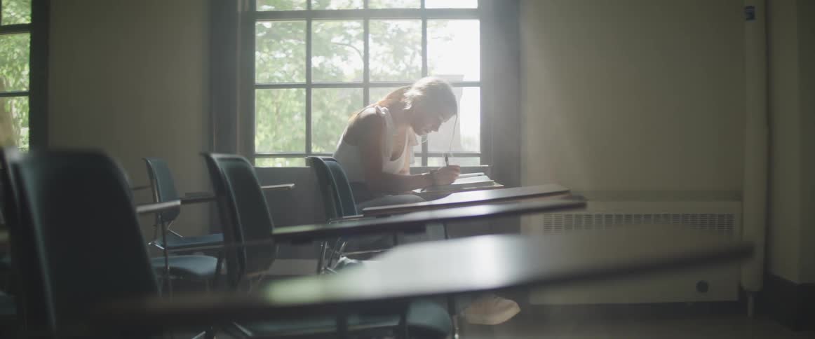 A teenager sits at a desk in a classroom and thinks and writes