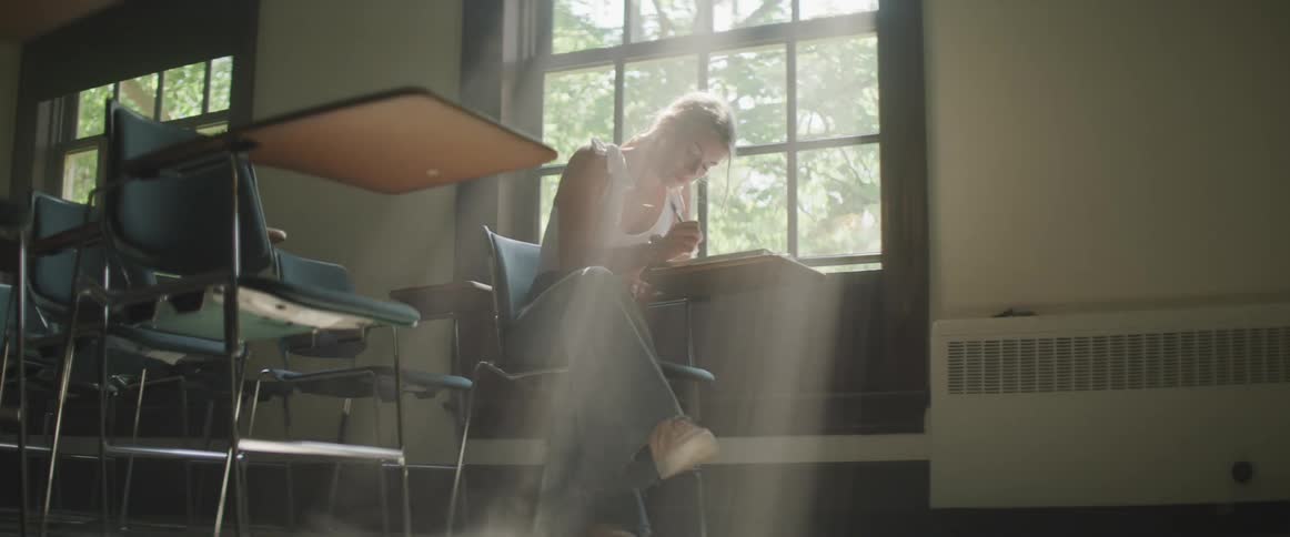 A woman sits at a school desk next to the window and starts to write after fidgeting