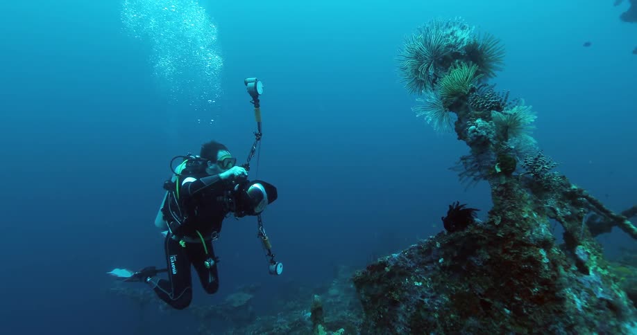 A scuba diver swims deep underwater while holding a camera in front of a reef